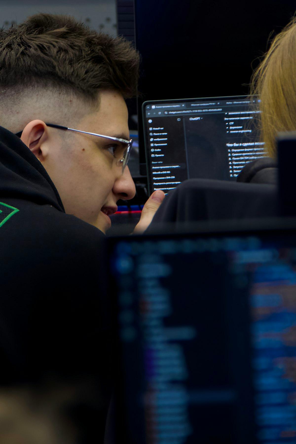 Estudiante de bachillerato con lentes, de perfil en un laboratorio de cómputo a oscuras, entre monitores con código y un portátil con un asistente de inteligencia artificial abierto