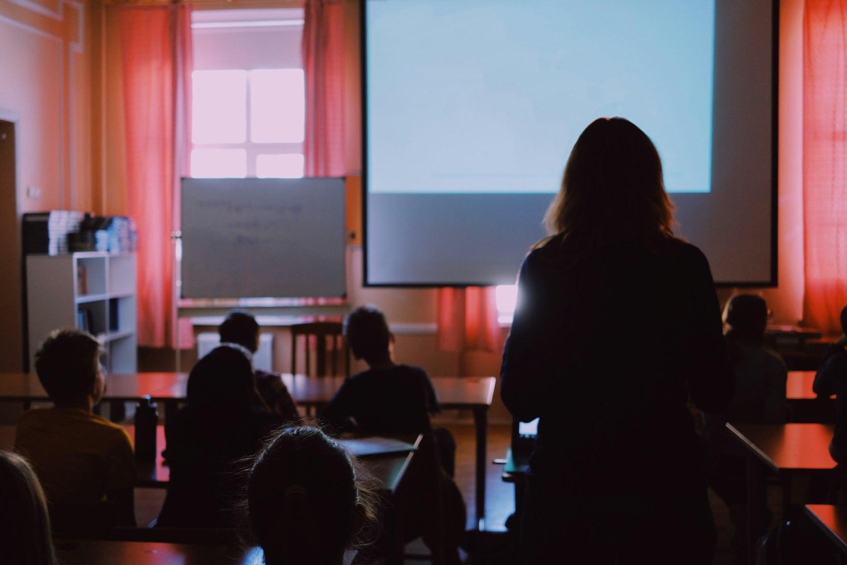 Aula a media luz con la clase frente a una pantalla encendida y la docente al frente